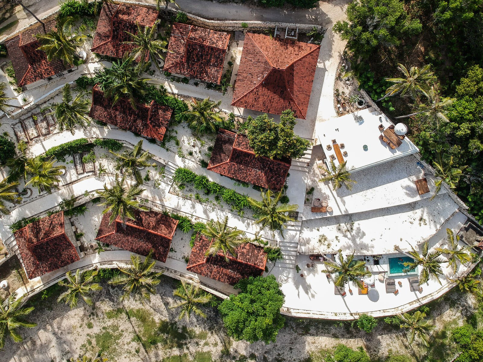 Aerial view of the whole Raja Lima village: wooden bungalows with red tile roofs, palm trees, white terraces and the plunge pools on the cliff