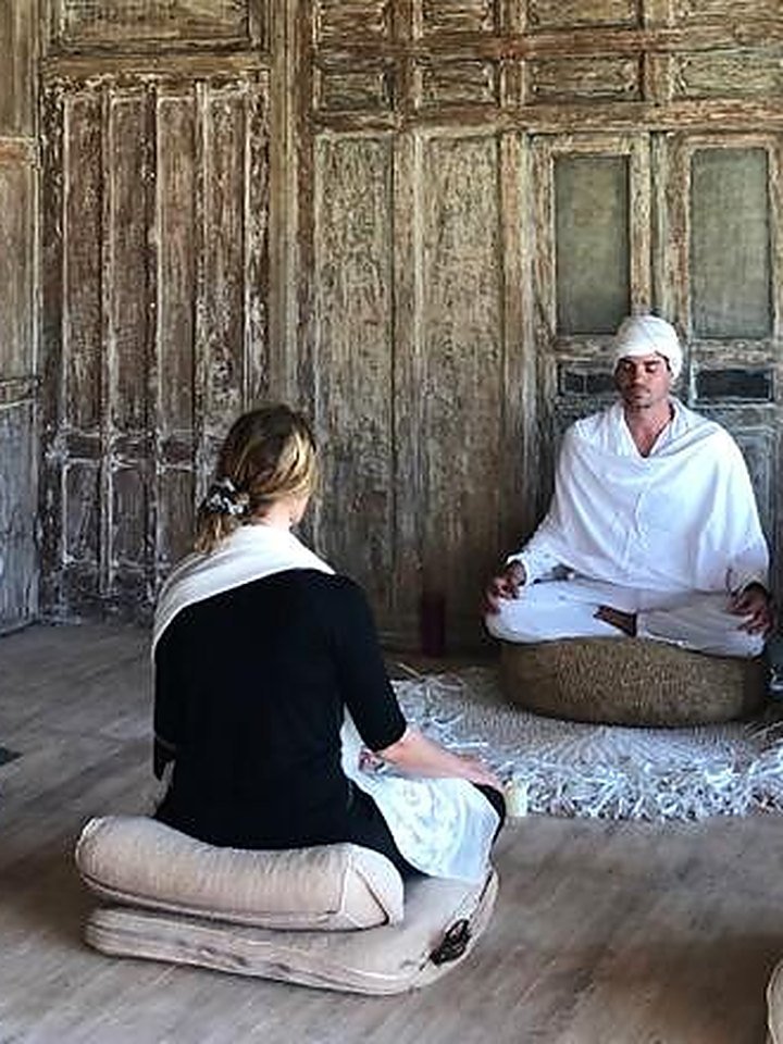 A guest meditating on a cushion facing the teacher in white in the wooden room