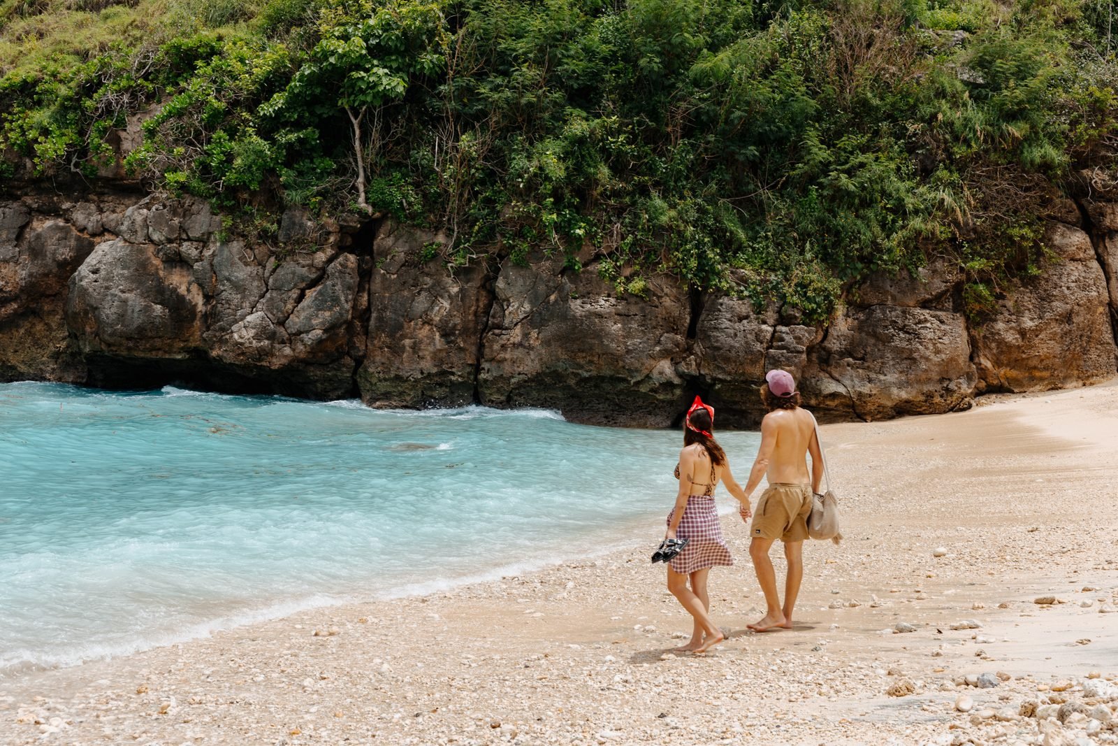 Two guests walking along the sand by the turquoise sea, below green cliffs
