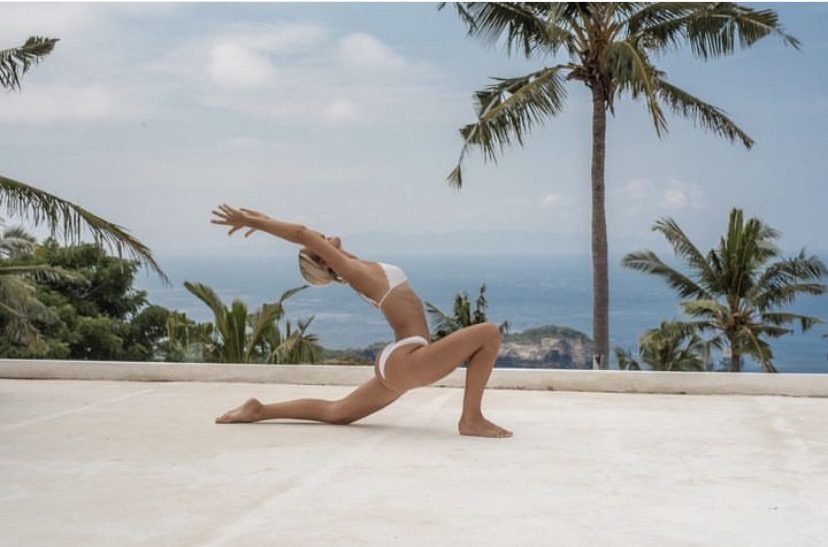 A guest in a low lunge yoga pose on the terrace, with palm trees and the ocean behind